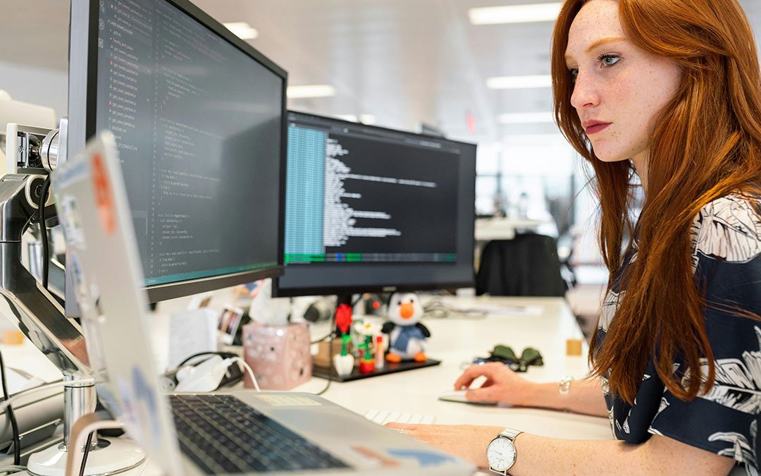 A developer at a desk watching code and a terminal across two monitors in an open-plan office